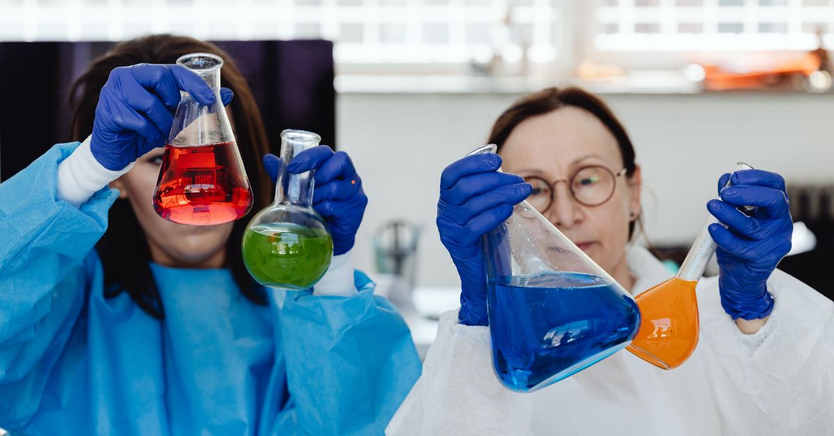 Two chemists, one male in a blue lab coat and one female in a white lab coat, both wearing protective gear while examining cleaning chemical samples